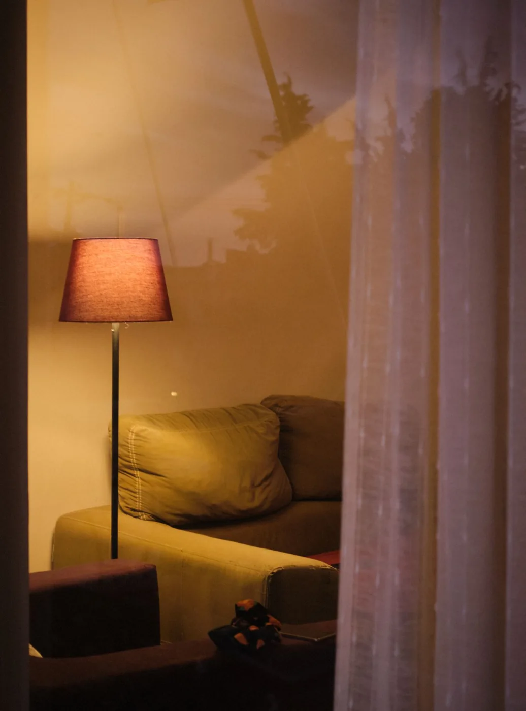 A glowing shaded lamp beside a sofa and pale curtains