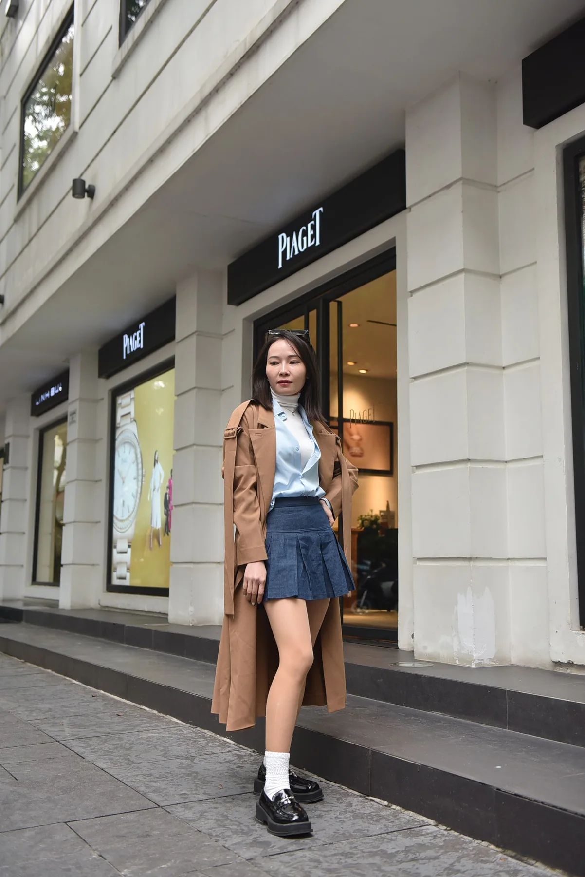 A woman wearing a brown long coat, pale shirt and blue pleated skirt outdoors
