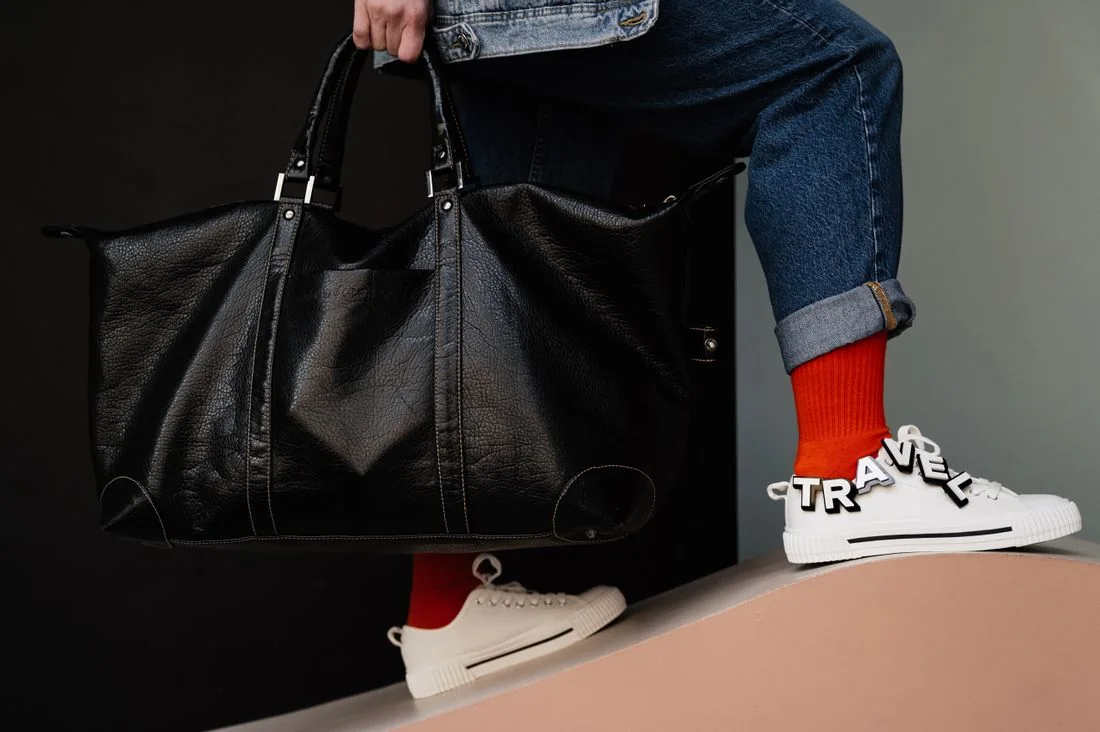 A person holding a dark duffel bag beside jeans and sneakers
